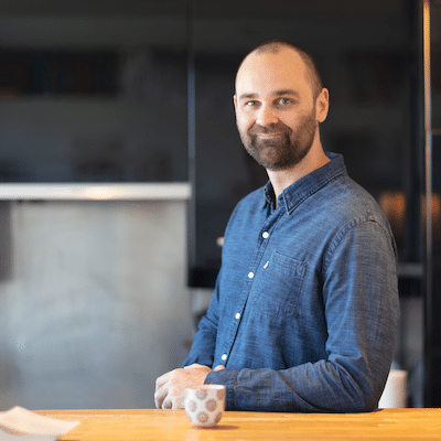 Man with a beard in a blue denim shirt standing at a wooden kitchen counter, smiling at the camera with a cup in front.